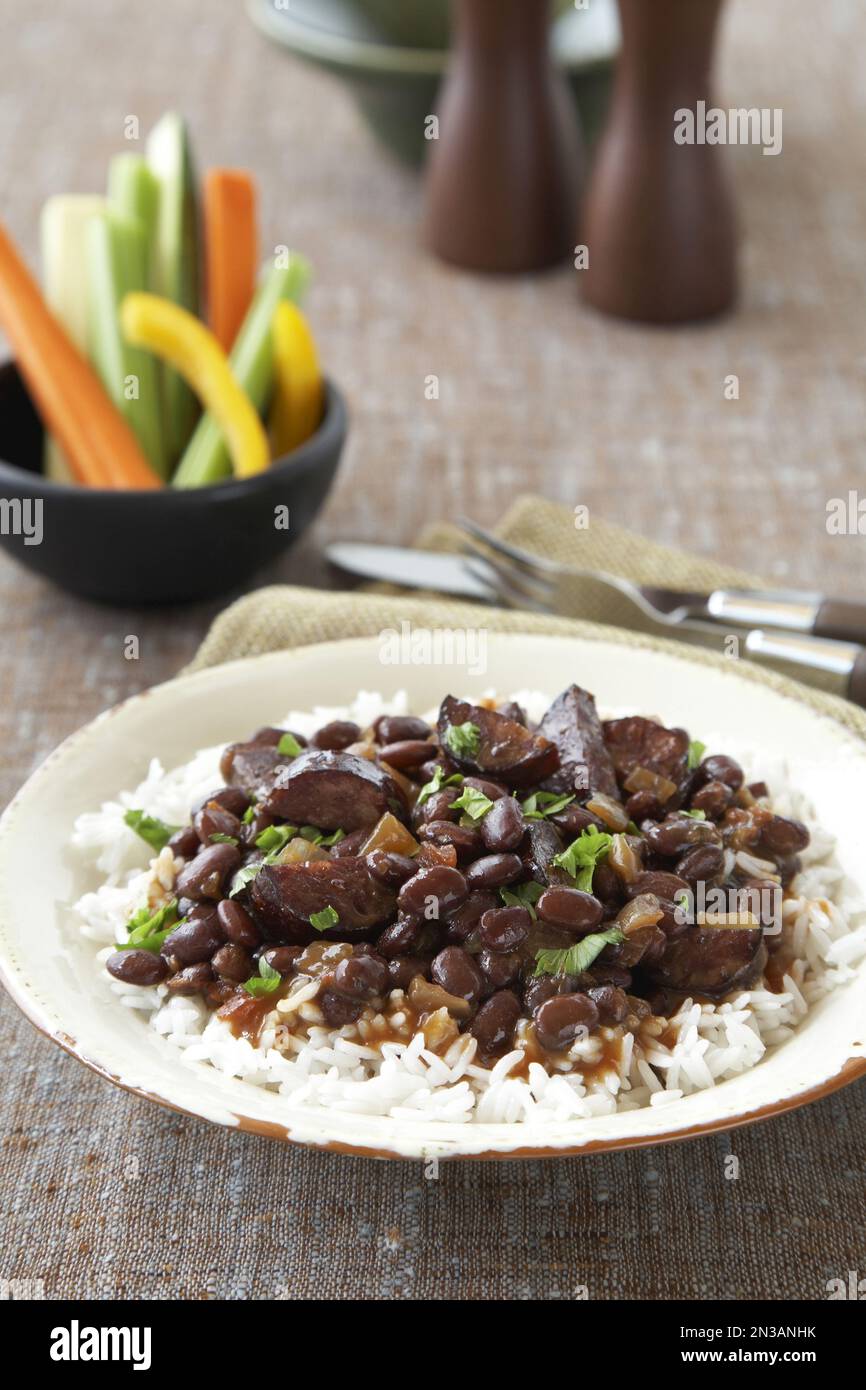 Schwarze Bohnen mit Wurst auf Reis, serviert mit rohen Gemüsestangen Stockfoto