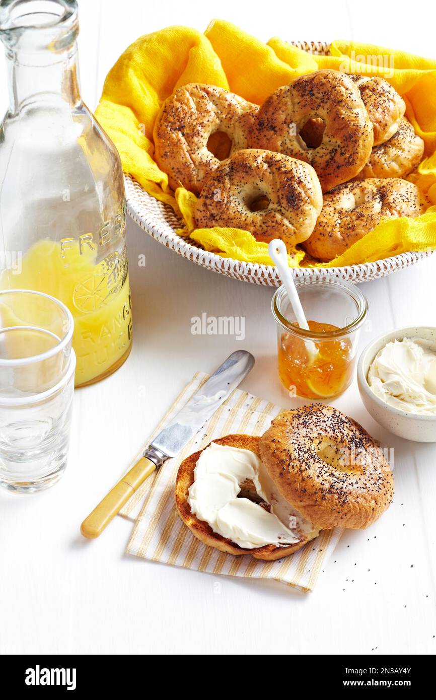 Bagel mit Frischkäse und Orangenmarmelade serviert mit frischem Orangensaft und einem Korb mit Bagels im Hintergrund. Stockfoto