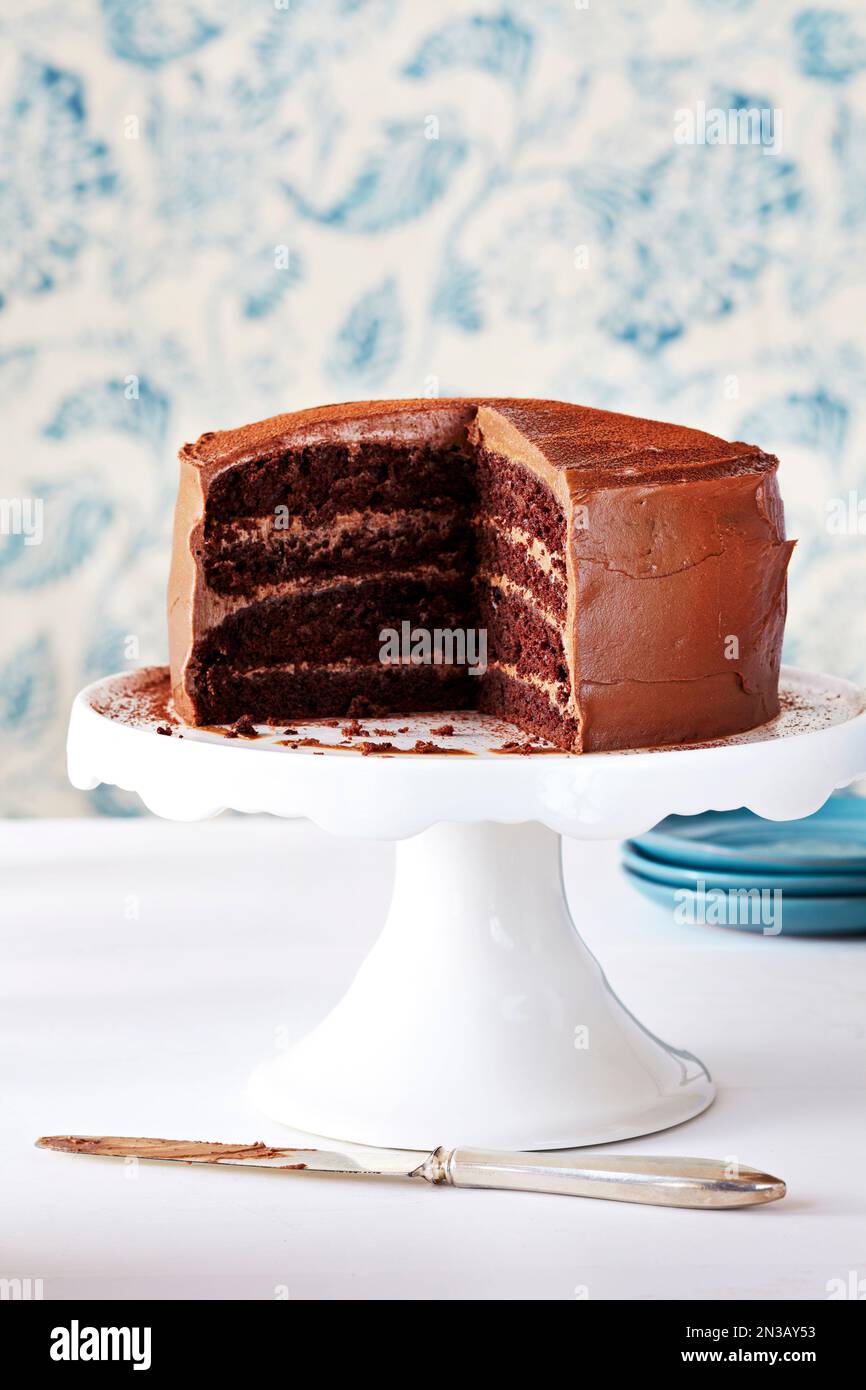 Schokoladenkuchen mit ein paar Scheiben auf einem weißen Kuchenständer mit weiß-blau gemustertem Hintergrund. Stockfoto