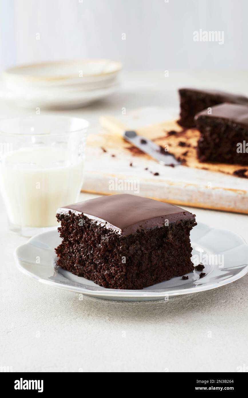 Ein Stück dunkler Schokoladenkuchen mit Schokoladenglasur auf einem Dessertteller mit einem Glas Milch Stockfoto