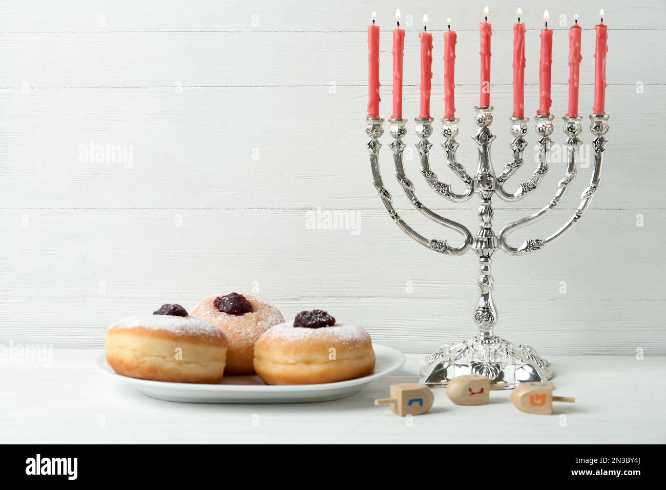 Silberne Menora, Dreidel mit He-, PE-, Nun-, Gimel- und Sufganiyot-Buchstaben auf weißem Holztisch, Platz für Text. Hanukka-Symbole Stockfoto