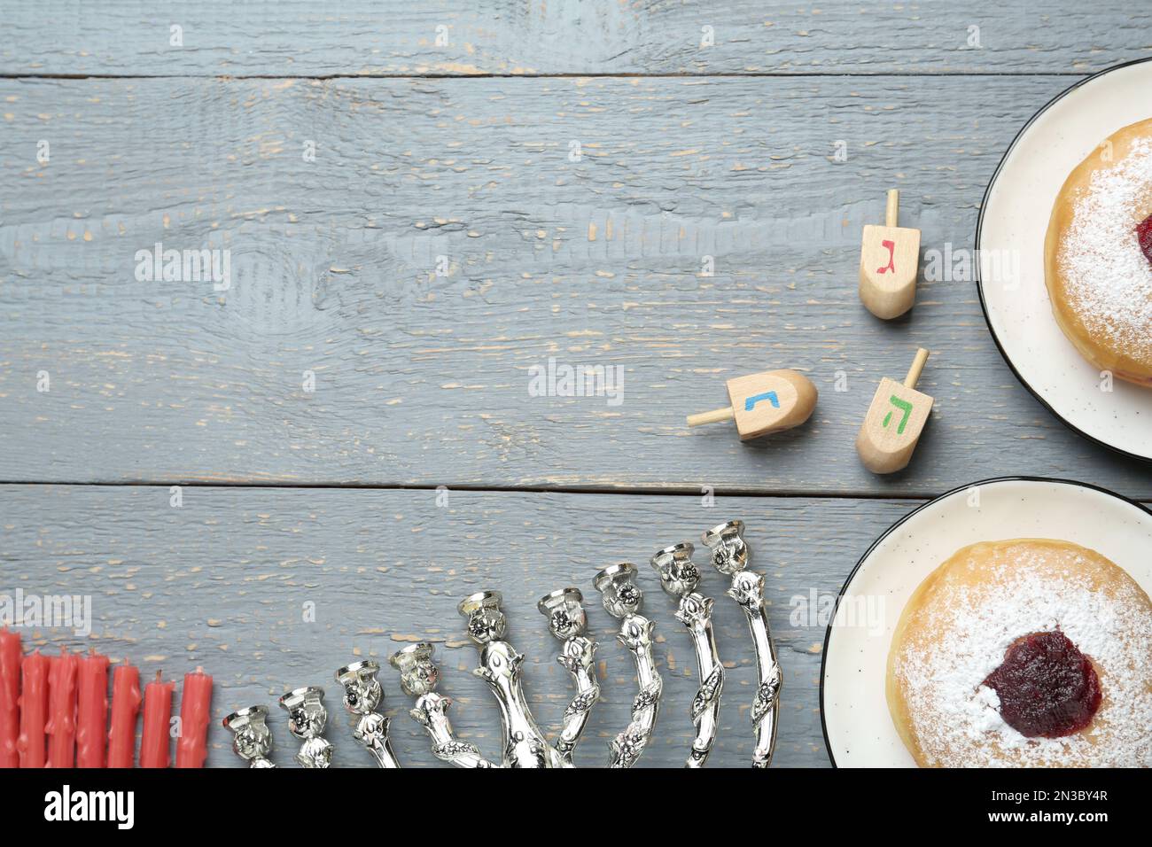 Flache Laienzusammensetzung aus Menorah, roten Kerzen, Dreidel mit He, PE, Nun, Gimel-Buchstaben und Sufganiyot auf grauem Holztisch, Platz für Text. Hanukka Stockfoto