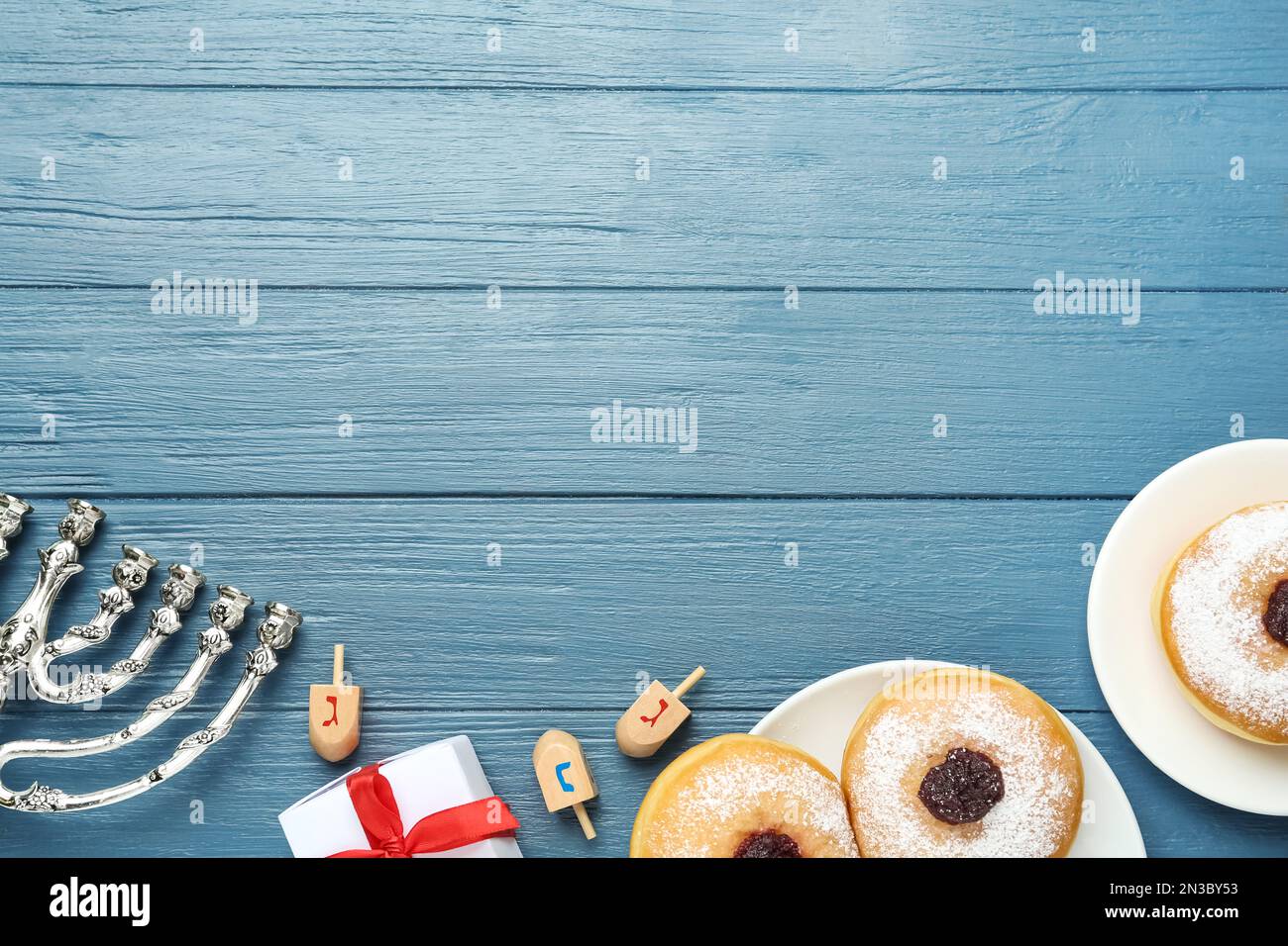 Flache Laienzusammensetzung von Menorah, Geschenkbox, Dreidel mit He, PE, Nun, Gimel-Buchstaben und Sufganiyot auf blauem Holztisch, Platz für Text. Hanukka-Symb Stockfoto