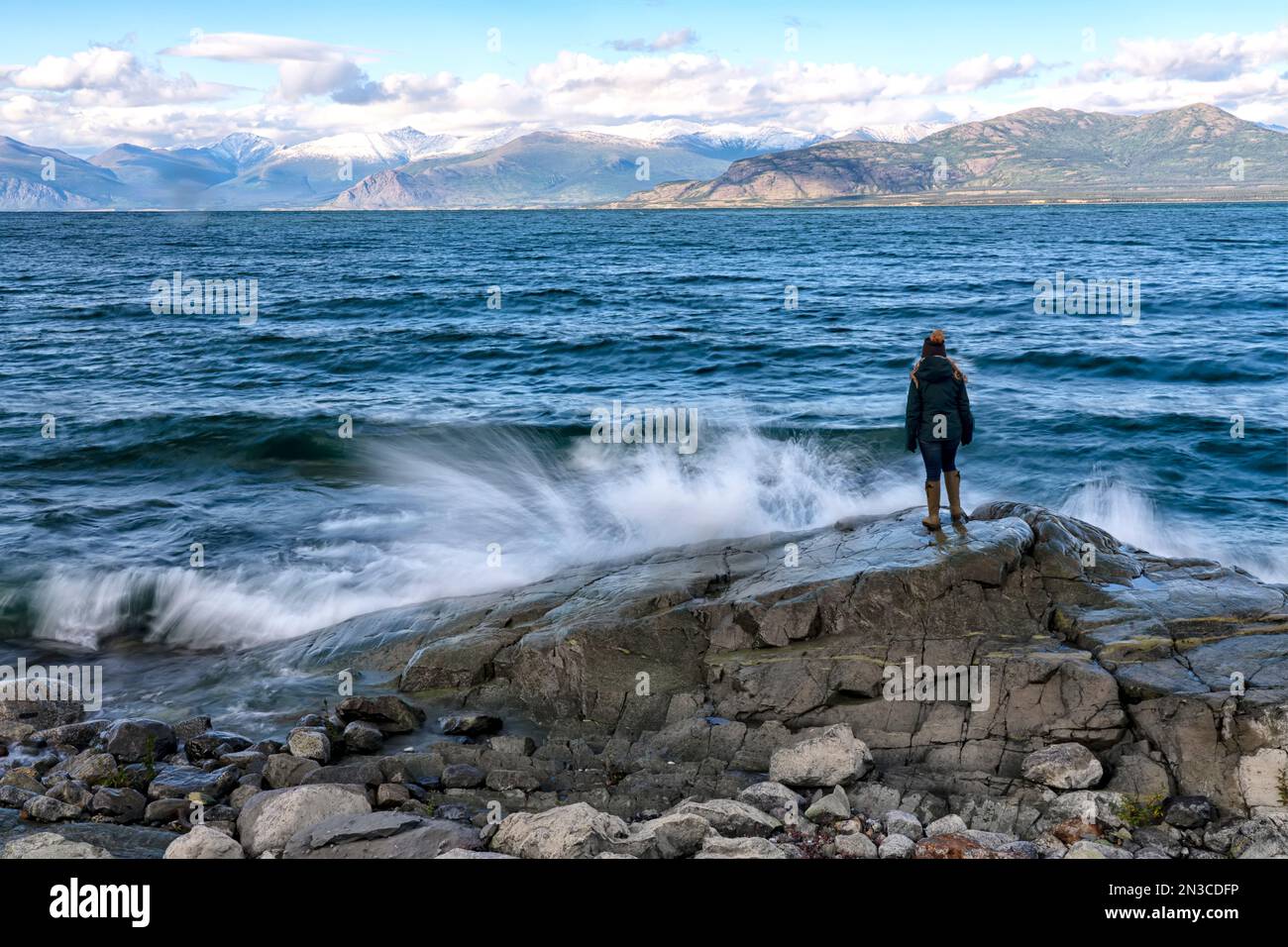 Blick von hinten auf eine Frau, die auf einem Felsen steht, während die Wellen an einem windigen Tag in das Ufer des Kluane Lake krachen; Yukon, Kanada Stockfoto