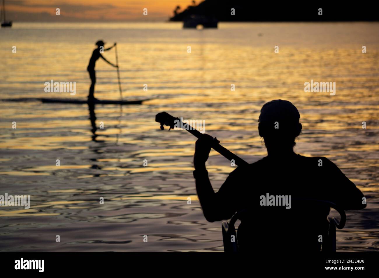 Mann, der auf einem Dock sitzt und bei Sonnenuntergang Banjo spielt, mit einem Stehpaddelboarder, der im Hintergrund vorbeipaddelt; Tahaa, Französisch-Polynesien Stockfoto