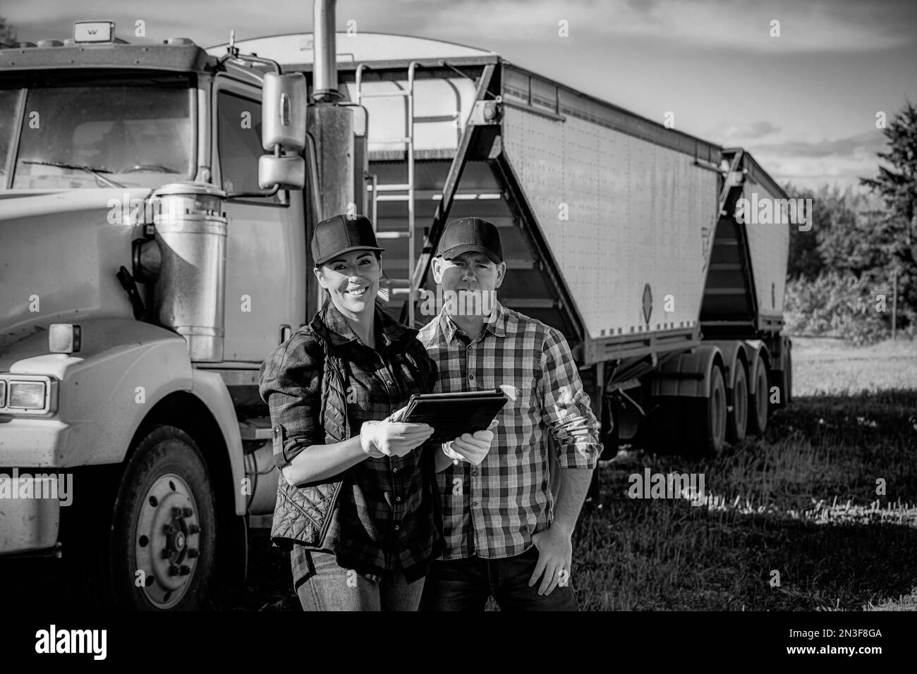 Porträt eines Ehemanns und einer Ehefrau, die ein tragbares drahtloses Gerät verwenden, um ihre Rapsernten zu verwalten und zu überwachen, während sie vor einem Traktor stehen... Stockfoto