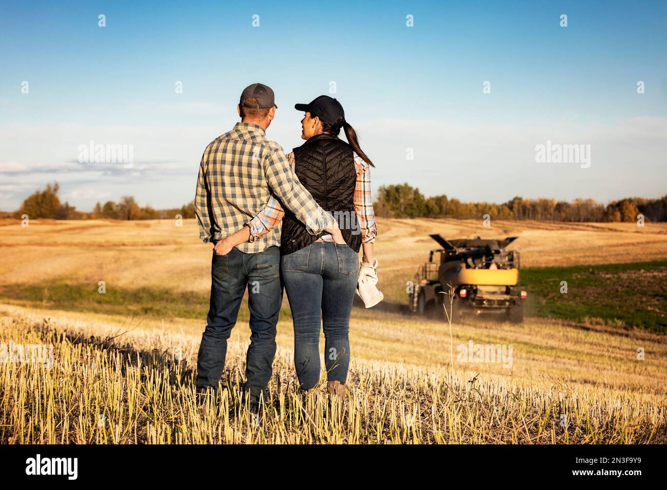 Ein Blick von hinten auf einen Mann und eine Frau, die auf einem Feld stehen und eine gute Zeit miteinander verbringen, während sie einen Mähdrescher beobachten, der ihren Herbst beendet... Stockfoto
