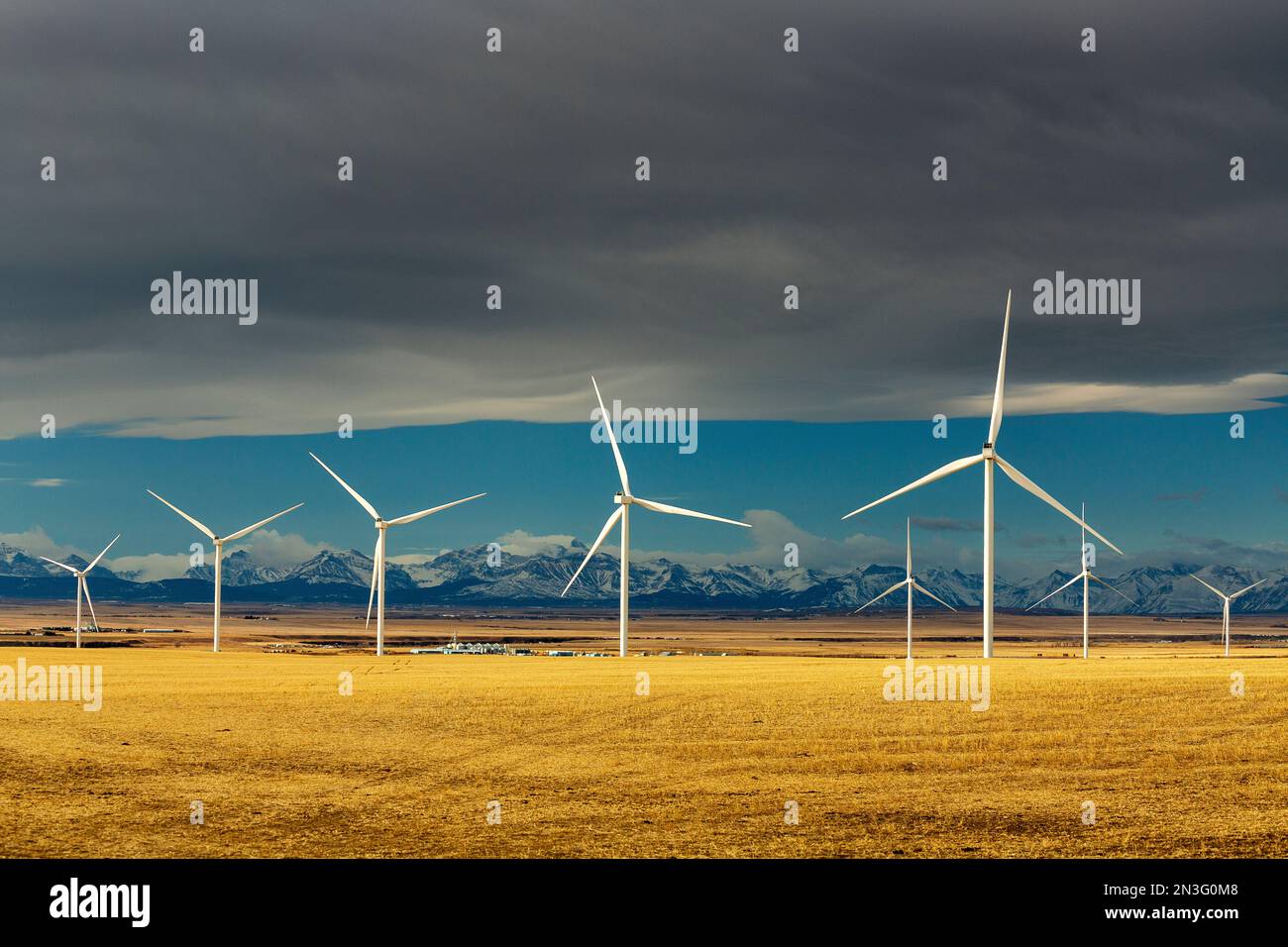 Große Windturbinen aus Metall auf einem Stoppelfeld mit Chinook-Bogen, blauem Himmel und Bergkette in der Ferne, nördlich von Glenwood, Alberta Stockfoto