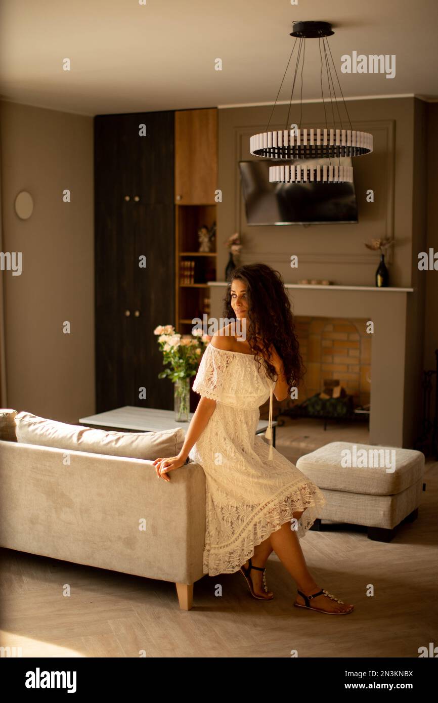 Hübsche junge Frau mit lockigem Haar entspannt sich in ihrem eigenen Zimmer, gekleidet in einem weißen fließenden Kleid Stockfoto