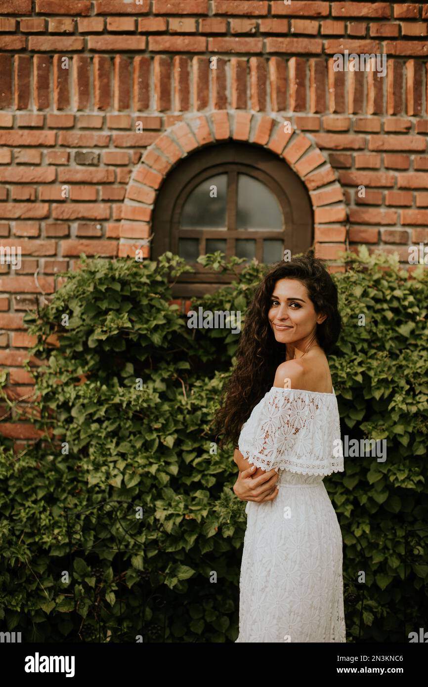 Hübsche junge Frau mit lockigem Haar steht in einem wunderschönen Garten, gekleidet in einem weißen Kleid. Sie sieht entspannt und zufrieden aus Stockfoto