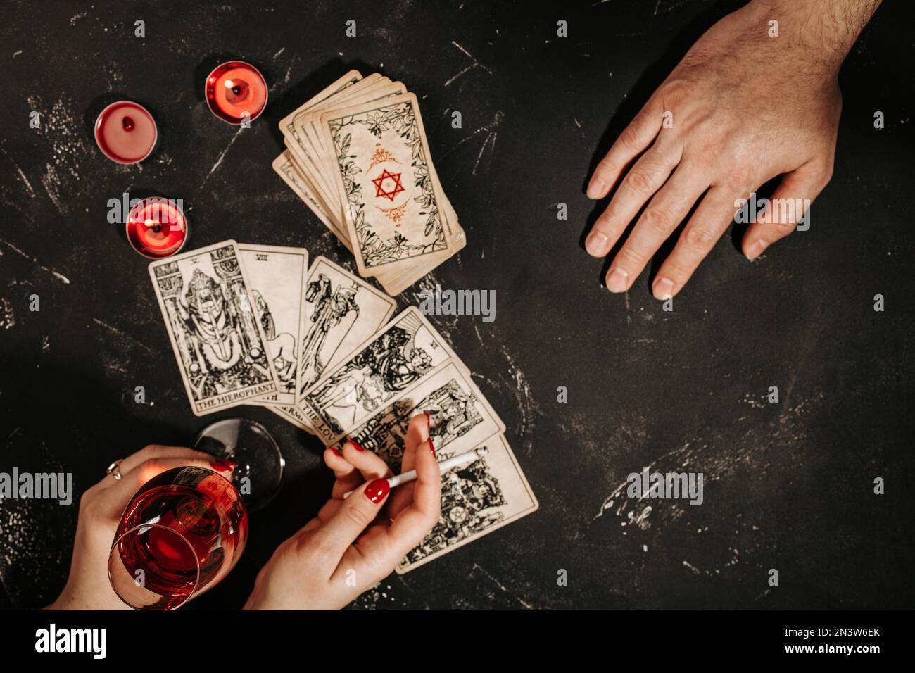 Weibliche Hände der Wahrsagerin mit einem Glas Rotwein beim Lesen von Tarot-Karten und männliche Hand des Besuchers auf einem schwarzen magischen Tisch mit rotem Hintergrund Stockfoto