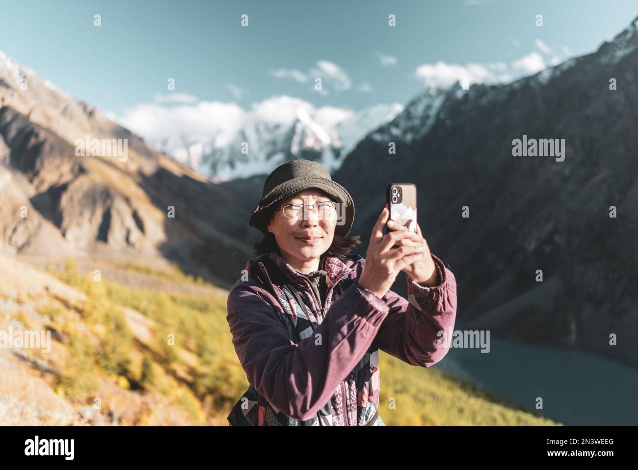 Ein asiatisches Mädchen mit Brille und Hut macht ein Selfie vor dem Hintergrund von Gletschern und schneebedeckten Bergen in der Nähe eines Sees in Altai. Stockfoto