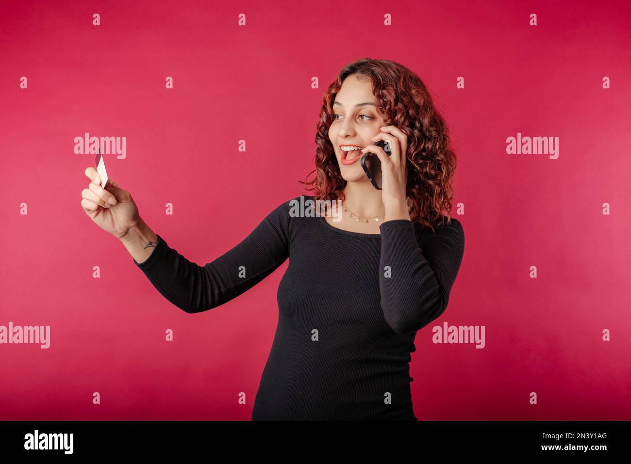 Porträt einer Frau, die isoliert auf rotem Hintergrund steht und die Nummer auf der Kreditkarte betrachtet und den Kauf telefonisch beim Kundendienst bestätigt. Stockfoto