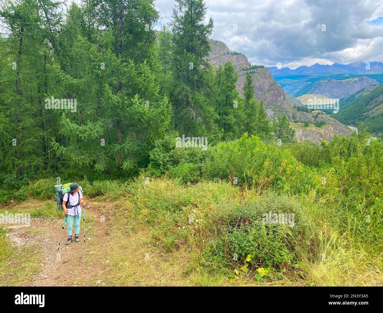 Ein Touristenmädchen mit einem Rucksack mit Brille und einem Hut ruht sich müde auf Wanderstöcken aus, vor dem Hintergrund des Panoramas des Altai Mo Stockfoto