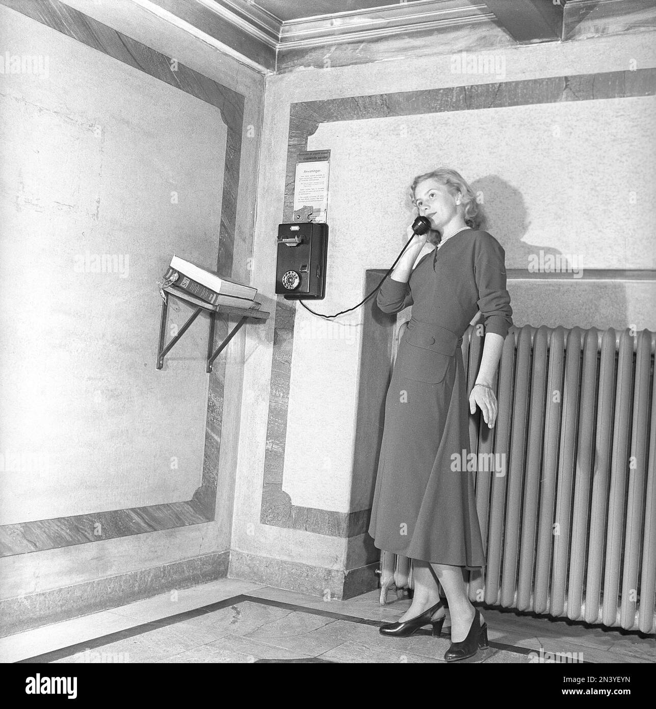 Damenmode in den 1940er. Eine junge Frau telefoniert mit einem Münztelefon. Sie steckten eine Münze und konnten die Nummer wählen und kurz reden. Neben ihr befinden sich die typischen Telefonverzeichnisse, in denen Sie die zu wählenden Nummern finden können, z. B. nach dem Nachnamen. Schweden 1949 Kristoffersson Ref AT37-10 Stockfoto