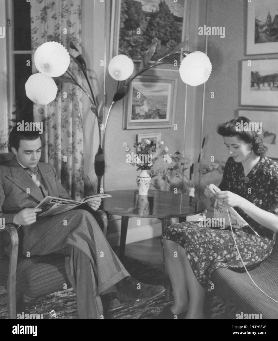 In den 1940er Jahren. Ein Paar zu Hause. Der Mann liest eine Zeitschrift, während sie strickt. Eine Lampe mit vier Globen ist im Fokus. Schweden 1946 Stockfoto