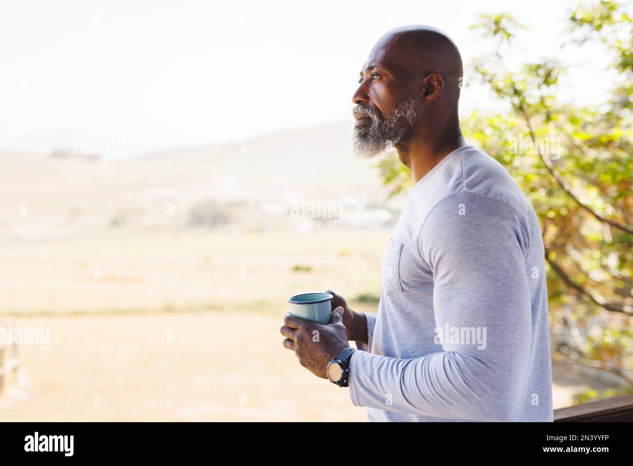 Kahlköpfiger afroamerikanischer Seniorenmann mit Tasse, der sich Gedanken macht und auf dem Balkon vor klarem Himmel steht Stockfoto