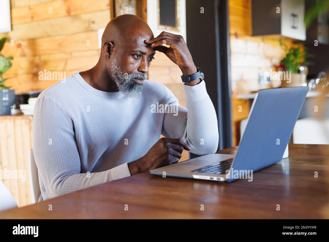 Gestresster, kahlköpfiger afroamerikanischer Senior-Mann mit Kopf in der Hand, der in einer Blockhütte auf einen Laptop schaut Stockfoto