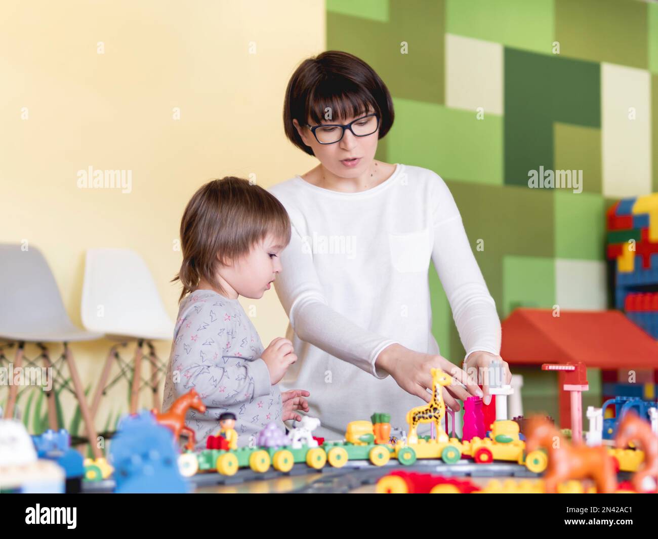 Ihr Kind spielt mit bunten Spielwürfeln, während seine Mutter oder sein Babysitter spielt. Der kleine Junge starrt auf den Spielzeugbauer. Das Innere des Kindergartens oder Kinderzimmers. Stockfoto