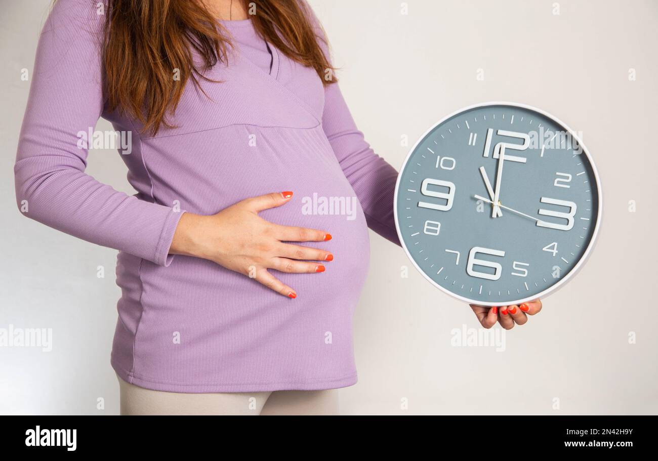 Ein schwangeres Mädchen im 3. Trimester hält eine Wanduhr vor dem Hintergrund eines großen Bauches. Der Begriff Frühgeburt, Vorstufen und To-Zeit Stockfoto