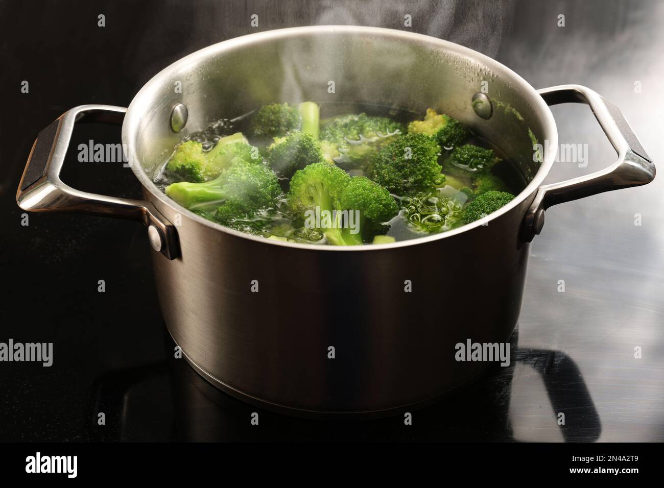 Brokkolistücke in einem Kochtopf aus Edelstahl mit kochendem Wasser und Dampf auf einem schwarzen Kochfeld, Zubereitung einer gesunden Gemüsesaal, Kopierraum, Sele Stockfoto