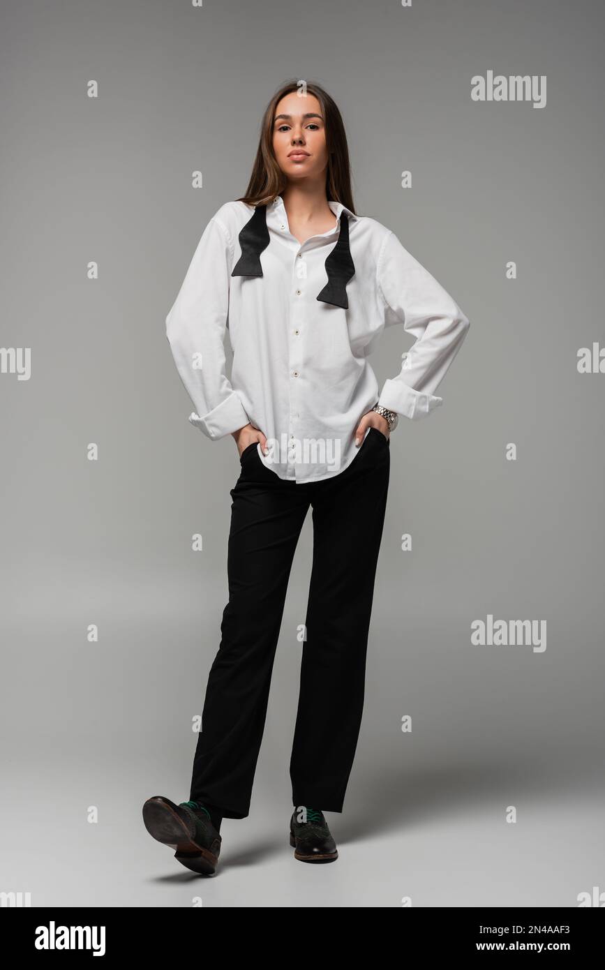 Lange, selbstbewusste Frau in weißem Hemd und Hose, die mit Händen in Taschen steht, auf grauem, Gleichstellungskonzept Stockfoto