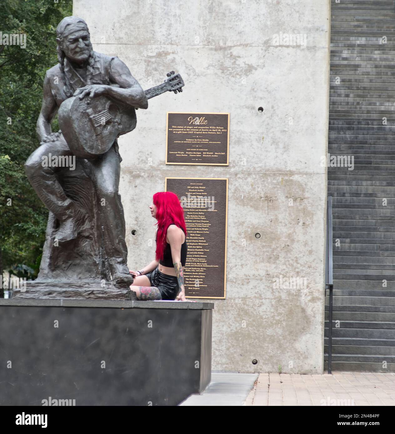 Ein Mädchen im Teenageralter mit gefärbtem magentafarbenem Haar und Ledershorts sitzt am Fuß der Willie Nelson Statue in Austin, Texas Stockfoto