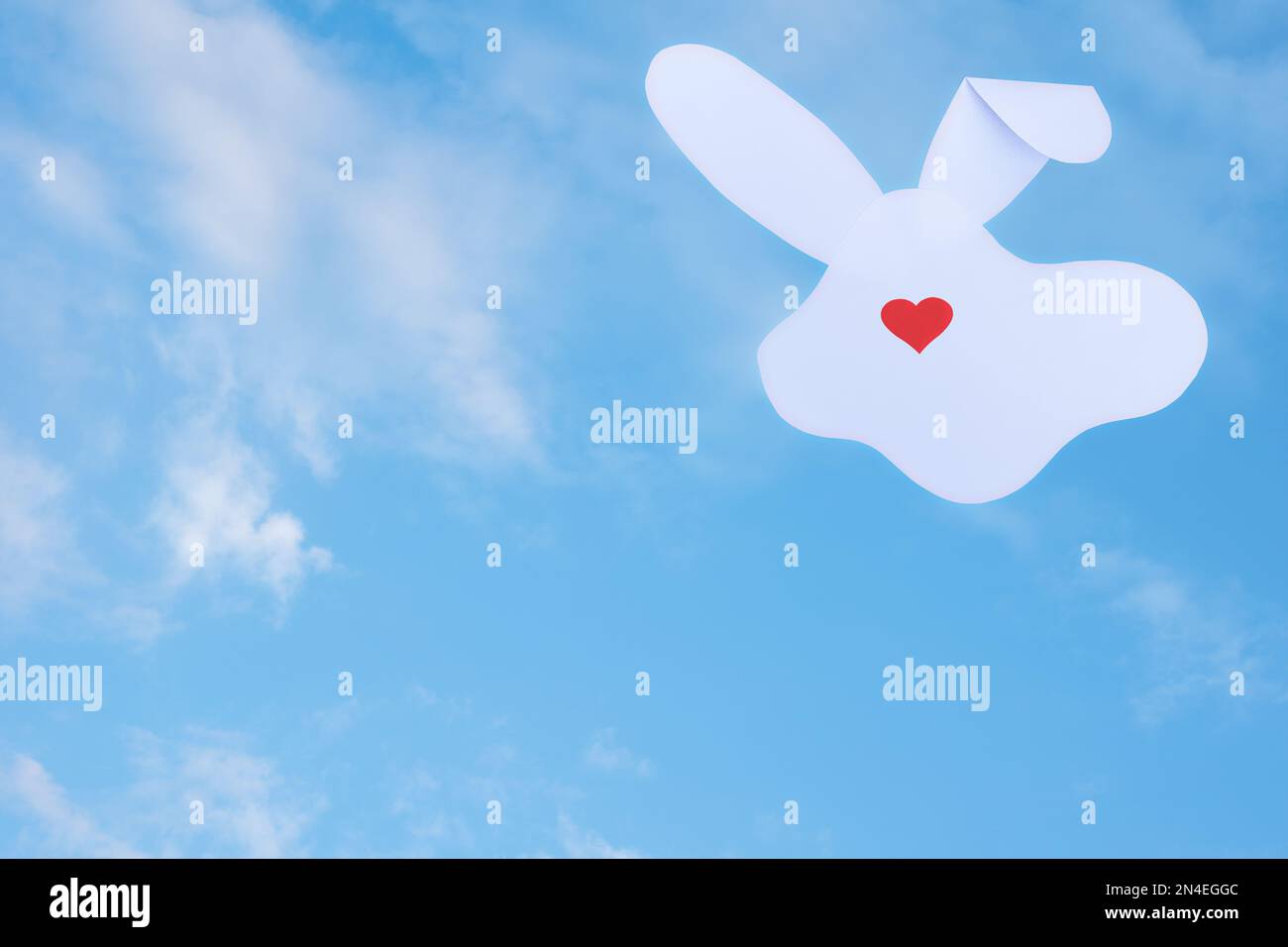 Kreative Karte aus einer Silhouette aus weißem Papierhasen und einem roten Herz anstelle einer Nase auf einem wolkigen Himmelshintergrund. Minimales Frühlingsferienkonzept Stockfoto