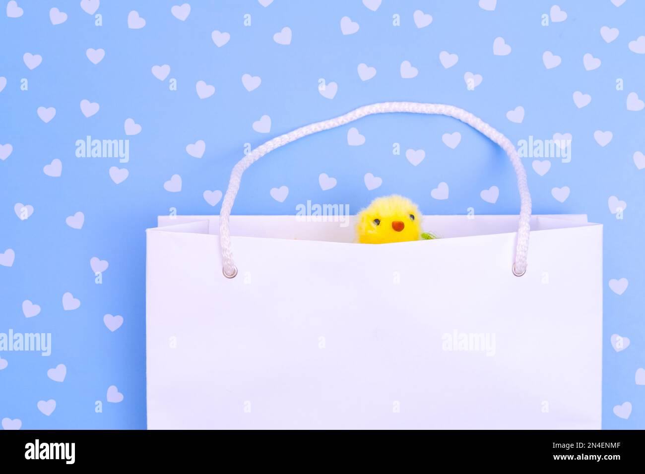 Weiße Einkaufstasche aus Papier mit gelbem, flauschigem Huhn auf blauem Hintergrund mit Herzen. Ostern einkaufen. Konzept Ostergeschenke, Vorbereitung, Feiertagssu Stockfoto