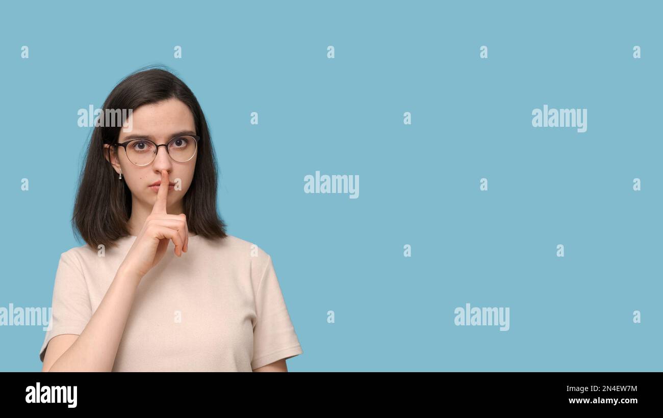 Porträt einer seriösen Schülerin mit Brille, die ihren Mund mit dem Finger auf blauem Hintergrund bedeckt, Banner. Das Mädchen will nicht reden. Keine Stockfoto