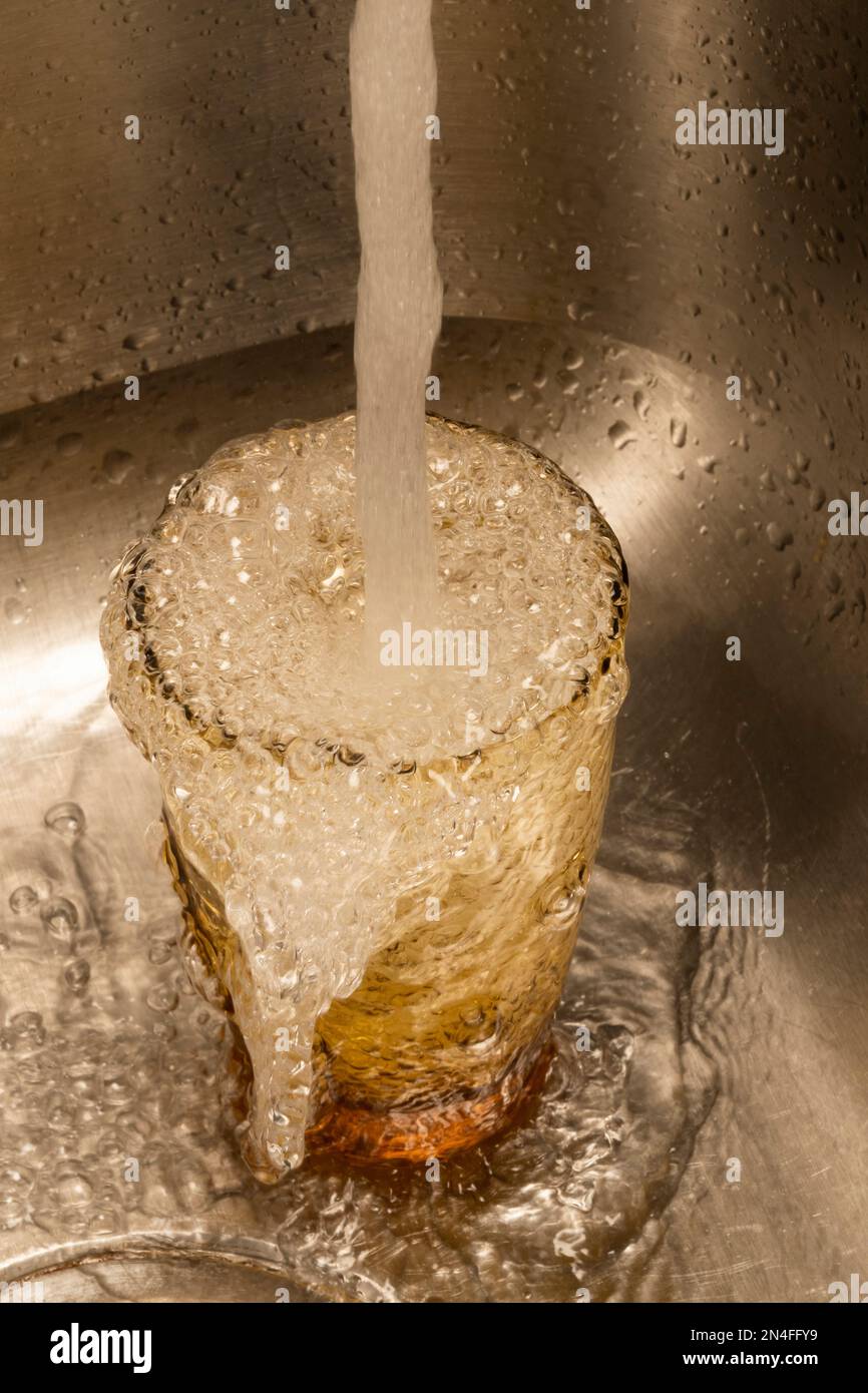 Vertikales Bild von Wasser, das über ein Trinkglas in einem Edelstahlwaschbecken fließt. Stockfoto