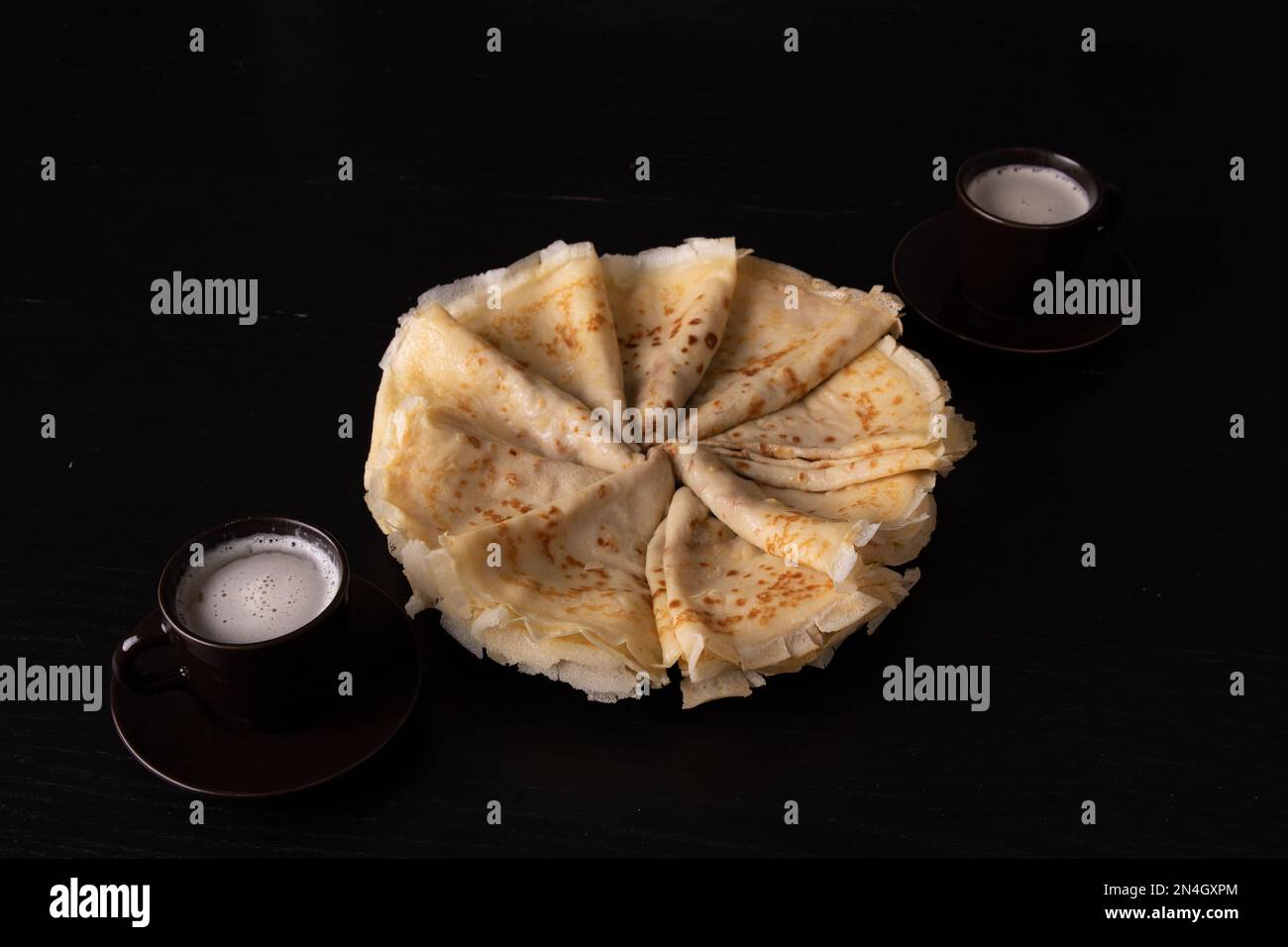 Foto-Pfannkuchen in einem Kreis auf einem Teller und zwei Tassen mit einem Getränk auf schwarzem Hintergrund Stockfoto