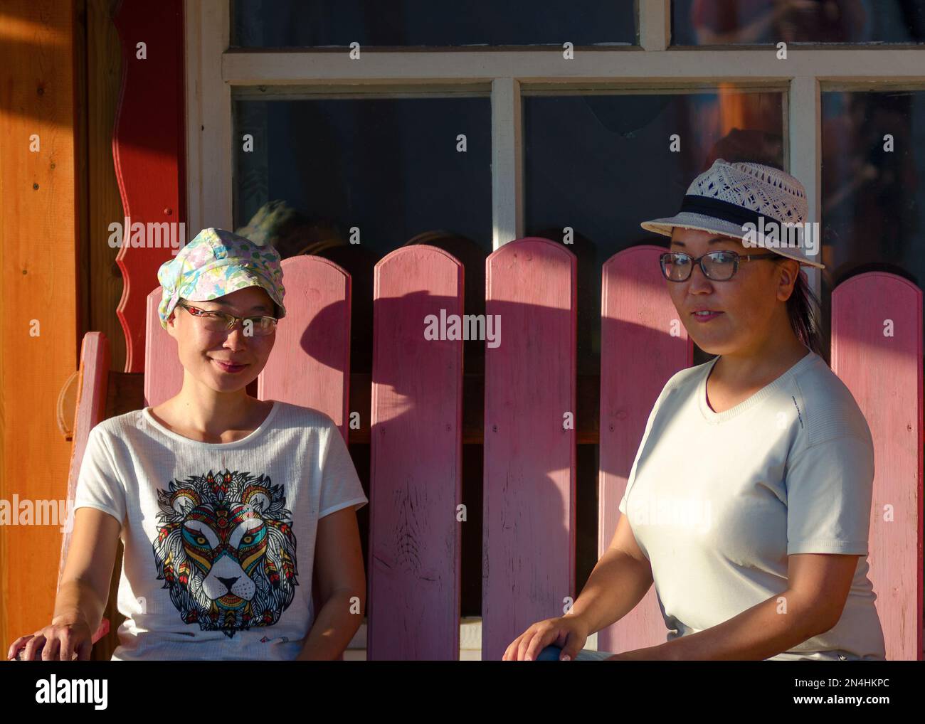 Zwei Mädchen Freundinnen Nachbarn Asiaten Yakuts posieren im Schatten auf einer Bank in der Sonne an einem Sommertag und lächeln. Stockfoto