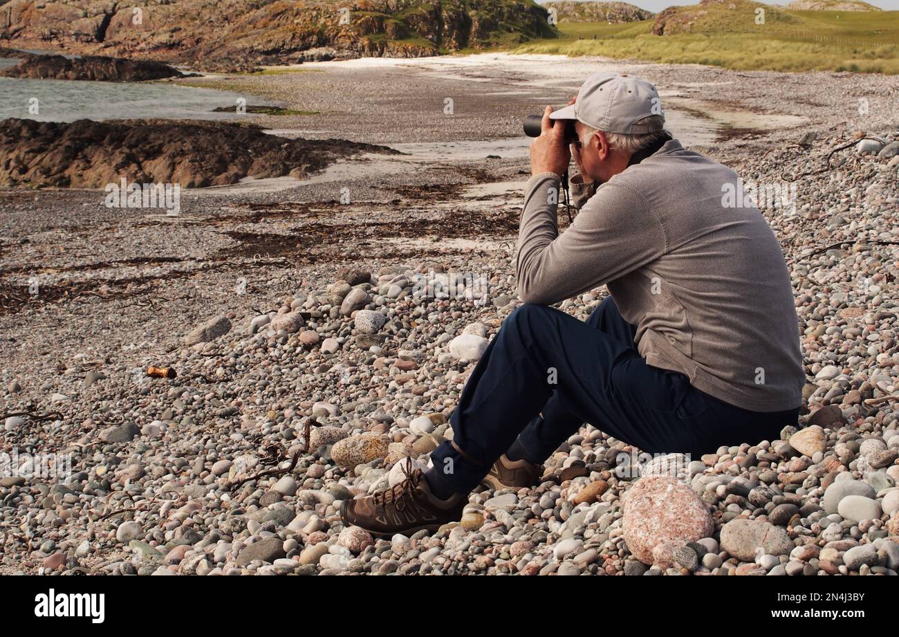 Mann 60+ sitzt an einem steinigen Strand Camas Cuil an t-Saimh, auf der Westseite von Iona, Schottland mit Blick auf das Meer durch Ferngläser Stockfoto