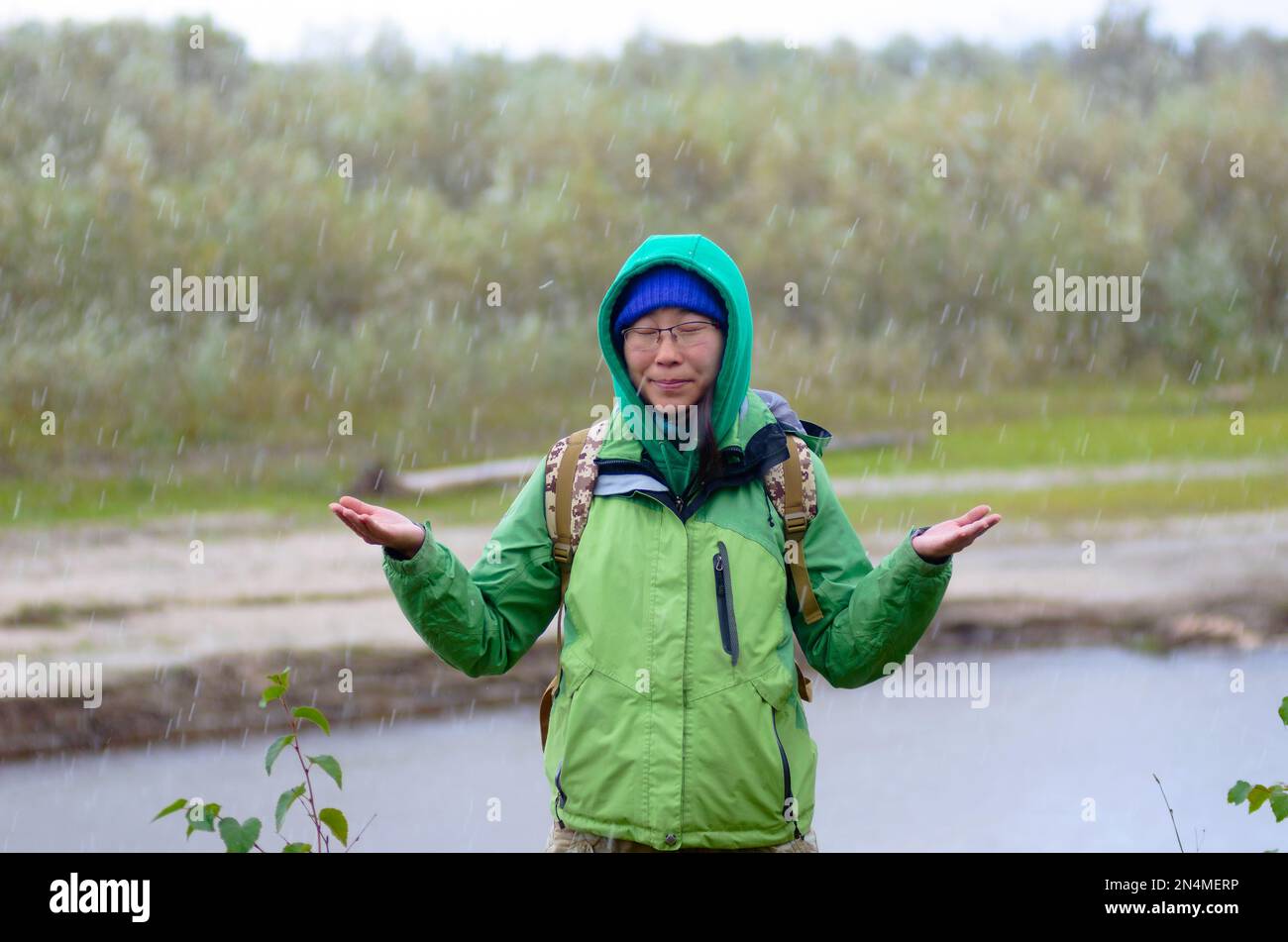 Yakut Asiatin Tourist in einer Jacke Hut und Kapuze und mit einem Rucksack schließt seine Augen, unter Hagel und Regen hält seine Hände gegen den Ba Stockfoto