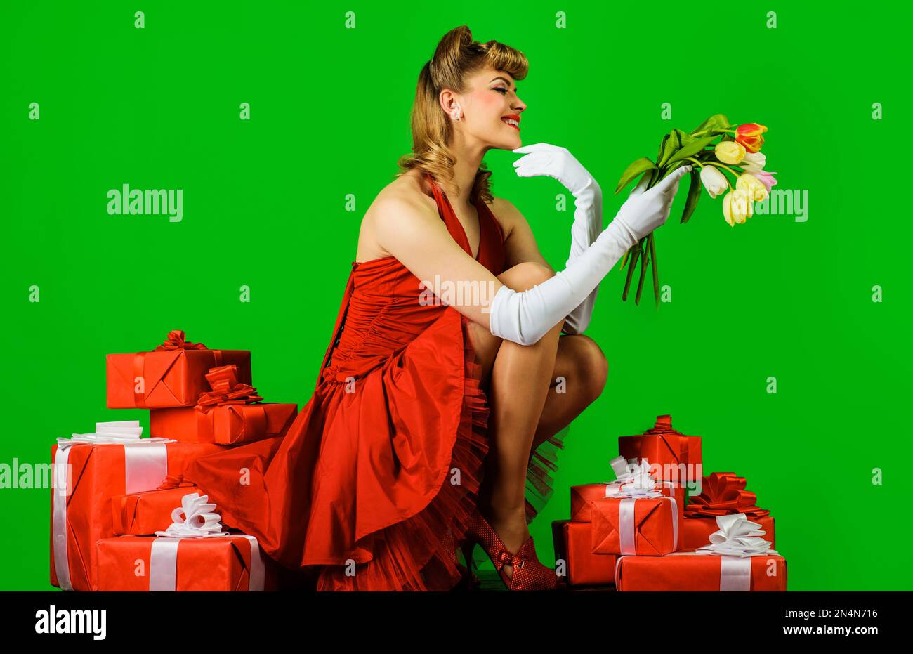 Lächelndes Mädchen mit Blumenstrauß und Geschenk. Frühlingsblumen und Geschenkbox. Wunderschöne Frau in rotem Kleid mit Retro-Frisur, Geschenkbox und Stockfoto