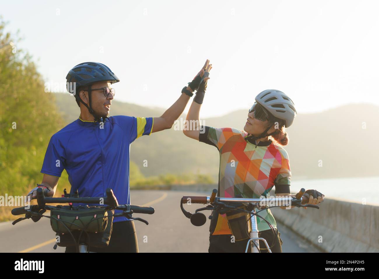 Das asiatische Radfahrer-Paar highfive fährt am Morgen mit dem Fahrrad zu ihrem Ziel um einen See mit wunderschönen Bergen im Hintergrund. Stockfoto