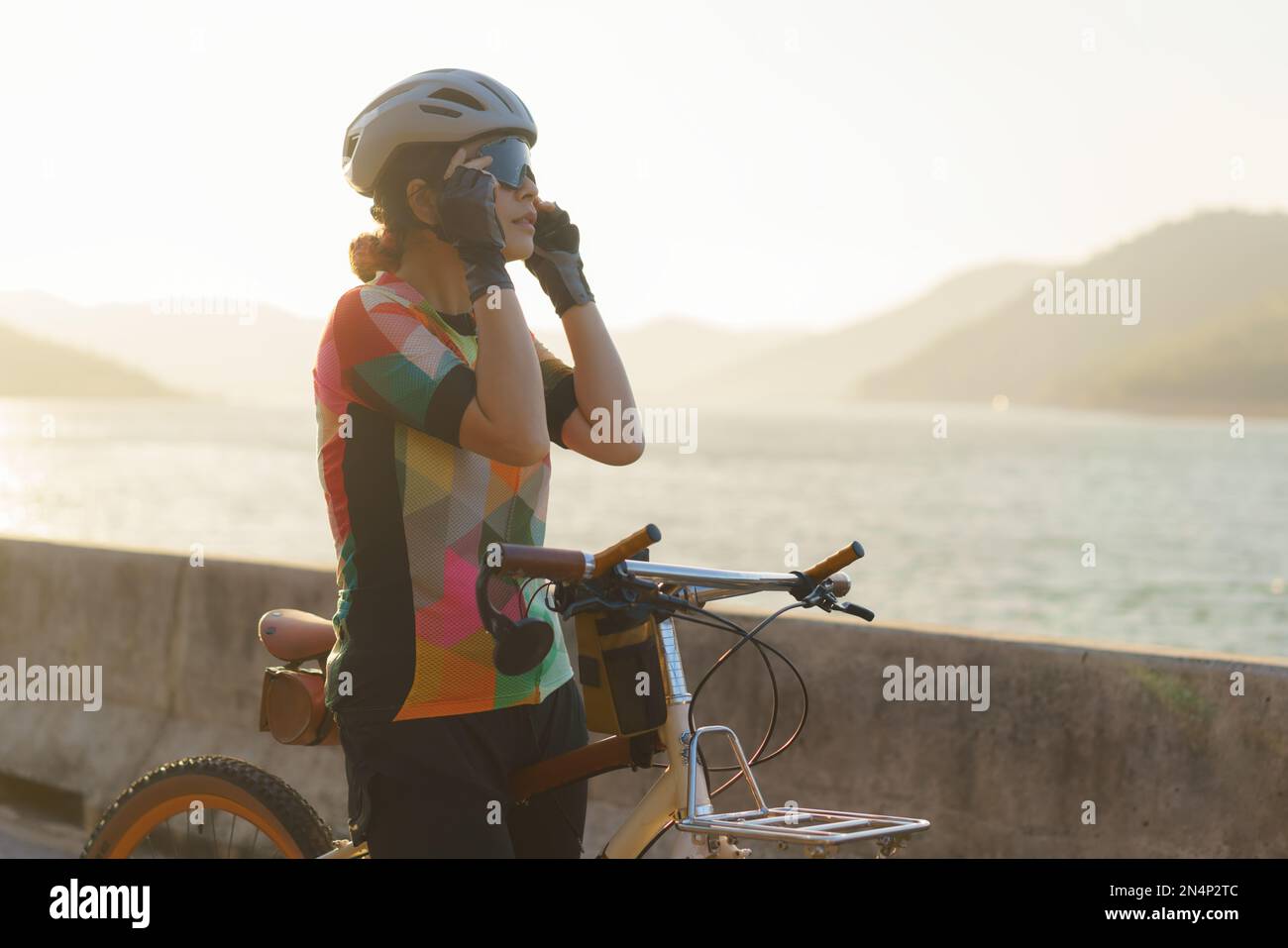 Eine asiatische Radfahrerin mit Sonnenbrille macht sich bereit für die morgendliche Radtour um den See mit wunderschönem Blick auf die Berge im Hintergrund. Stockfoto