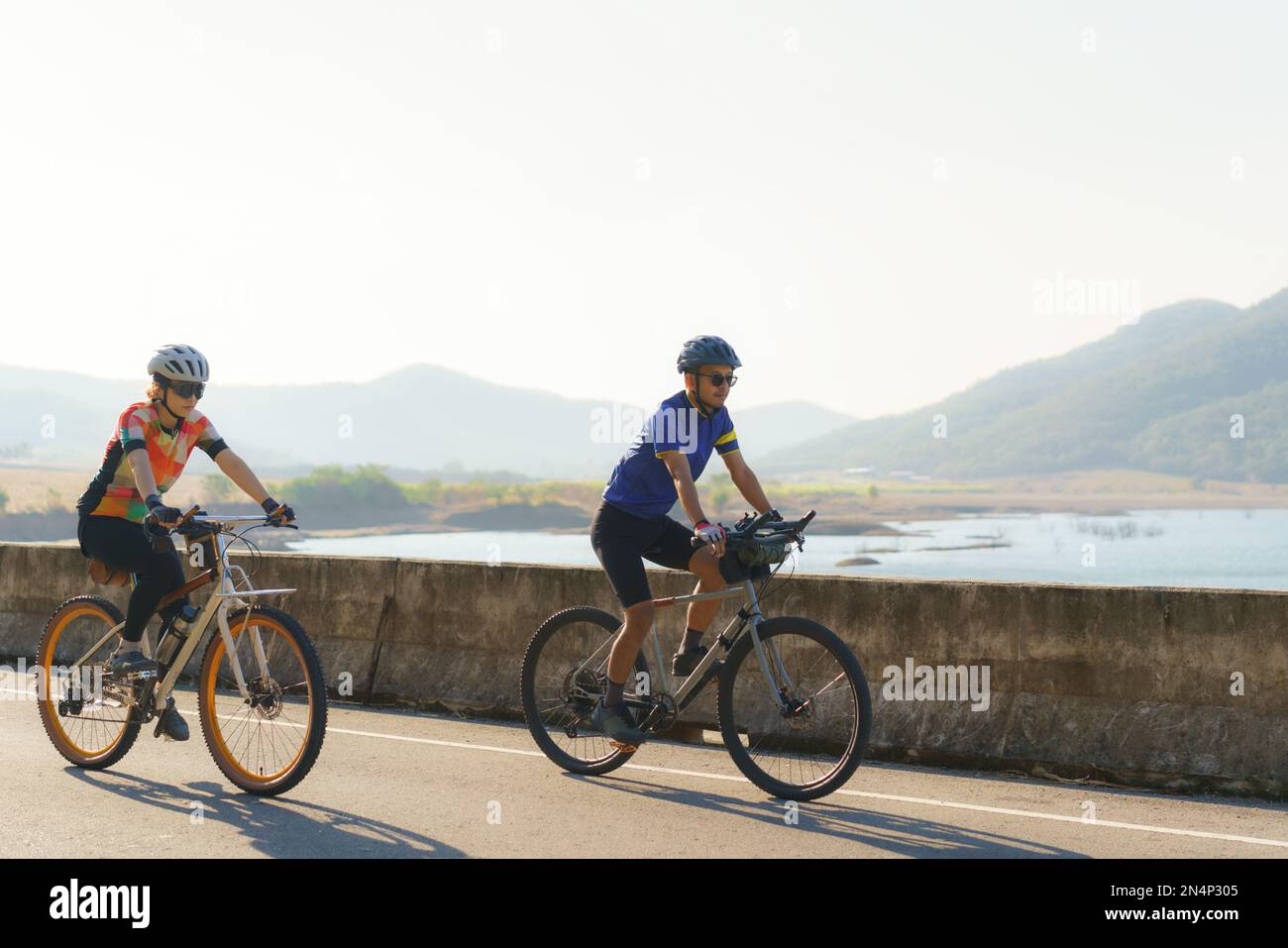 Asiatisches Fahrradpaar, das am Morgen gemeinsam um den See reitet, mit wunderschönem Blick auf die Berge im Hintergrund. Stockfoto