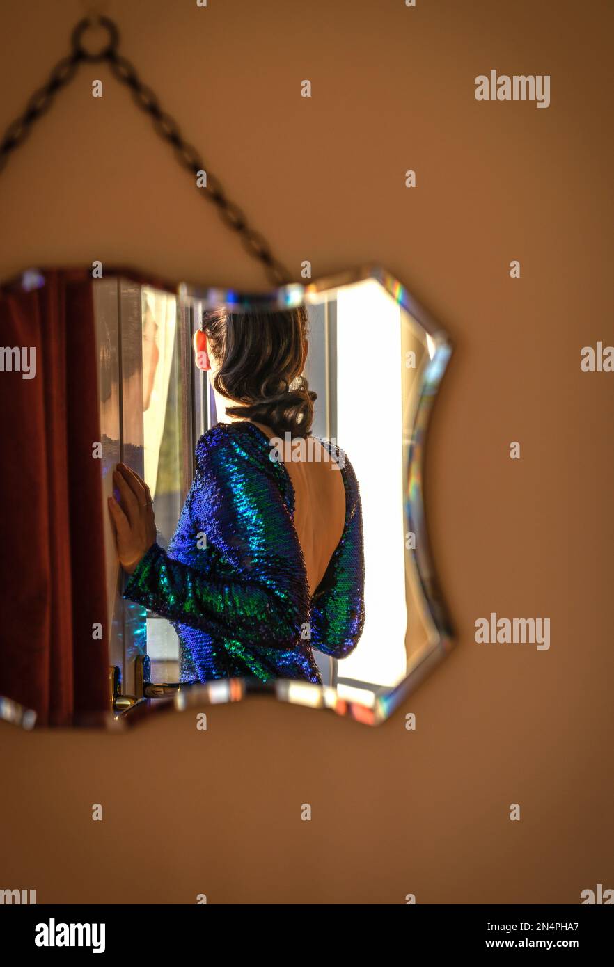 Reflexion in einem wandmontierten Spiegel einer Frau von hinten in einem blau-grünen, offenen Partykleid mit Pailletten Stockfoto