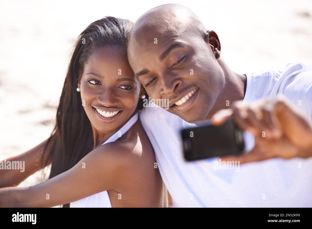 Lächle für mich... Ein glückliches, junges afrikanisches Paar, das lächelt, während es am Strand einen Schnappschuss macht. Stockfoto