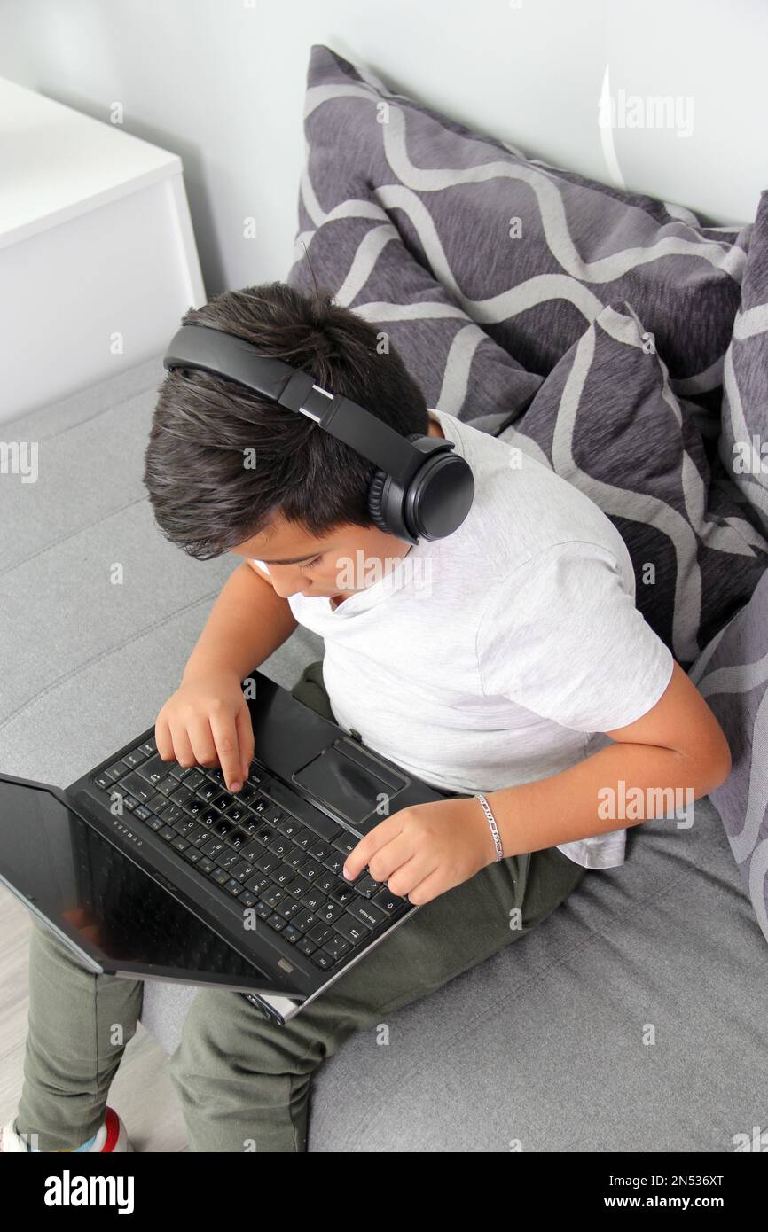 Der 9-jährige hispanische Junge spielt Videospiele auf seinem Laptop mit Kopfhörern, was zu Übergewicht und schlechter Sitzhaltung führt Stockfoto