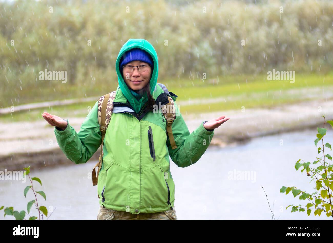 Yakut Asiatin Tourist in einer Jacke mit Hut und Kapuze und mit einem Rucksack lächelt, Hagel und Regen halten seine Hände vor dem Hintergrund des riv Stockfoto