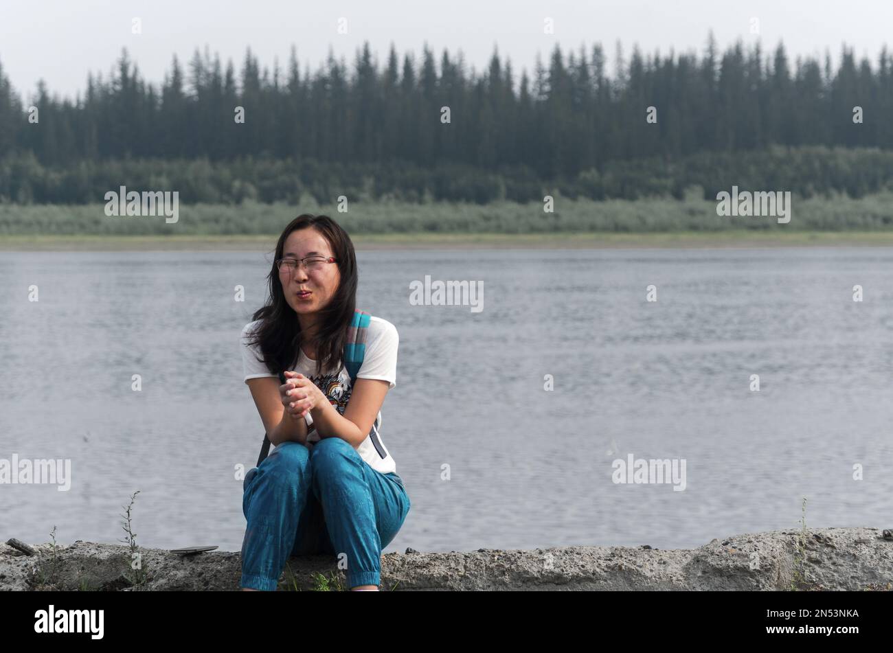 Überraschter Yakut Asiate mit Brille, junges Mädchen, das auf dem alten Pier des Flusses Viluy in den nördlichen Taiga-Tannen mit seiner Hand im Schloss sitzt Stockfoto