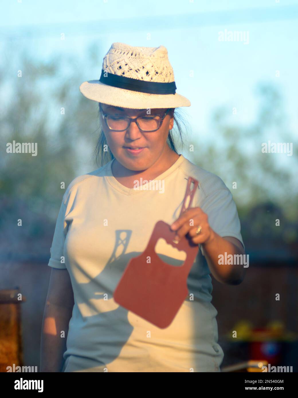 Yakut-Asiatin in Brille und Hut, die mit einem Feuerventilator im Grill wedelt, in den Strahlen der untergehenden Sonne. Stockfoto