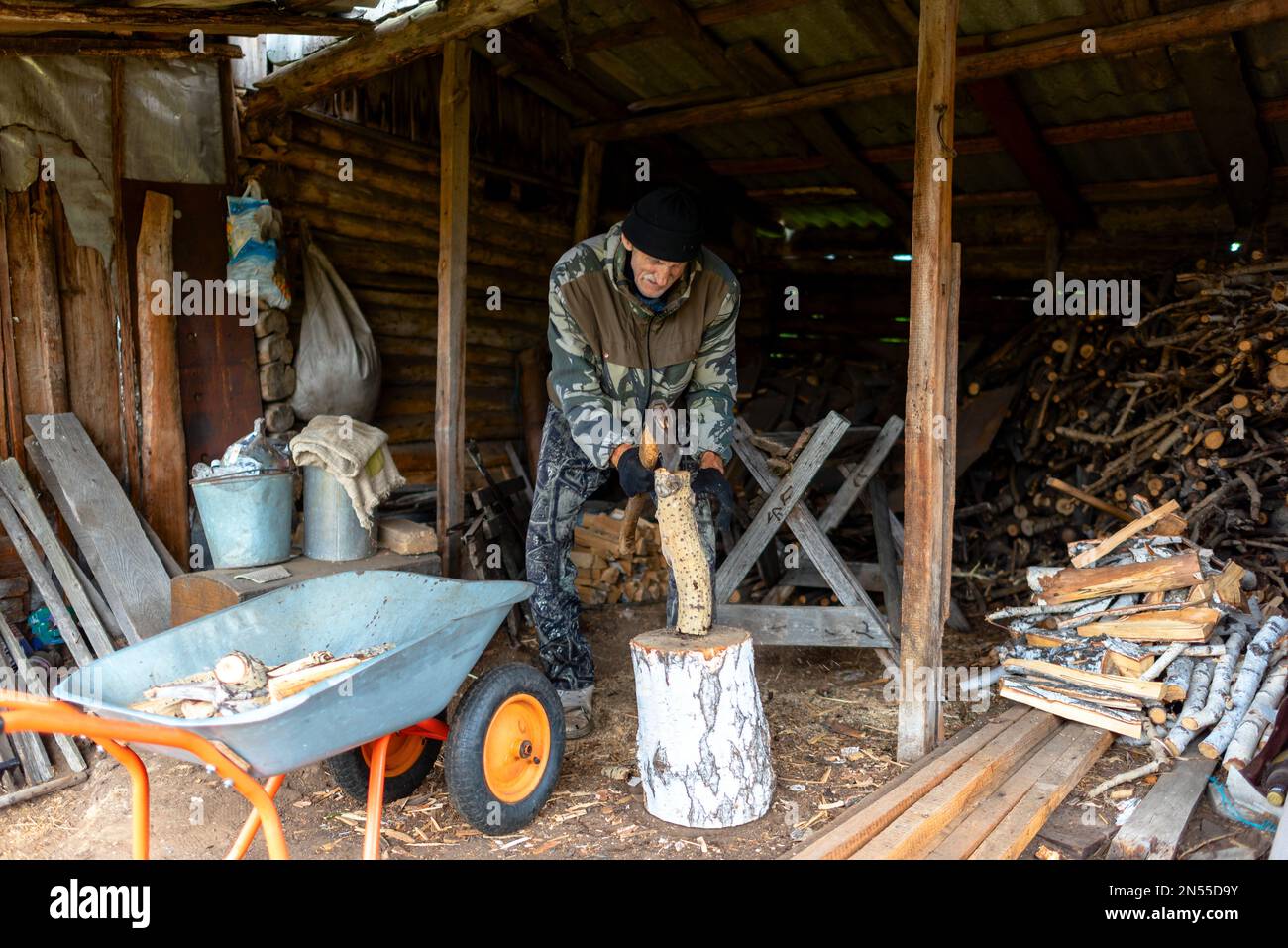 Ein älterer Russe hackte ermüdend Holz mit einer Axt in einer Scheune im Dorf. Stockfoto