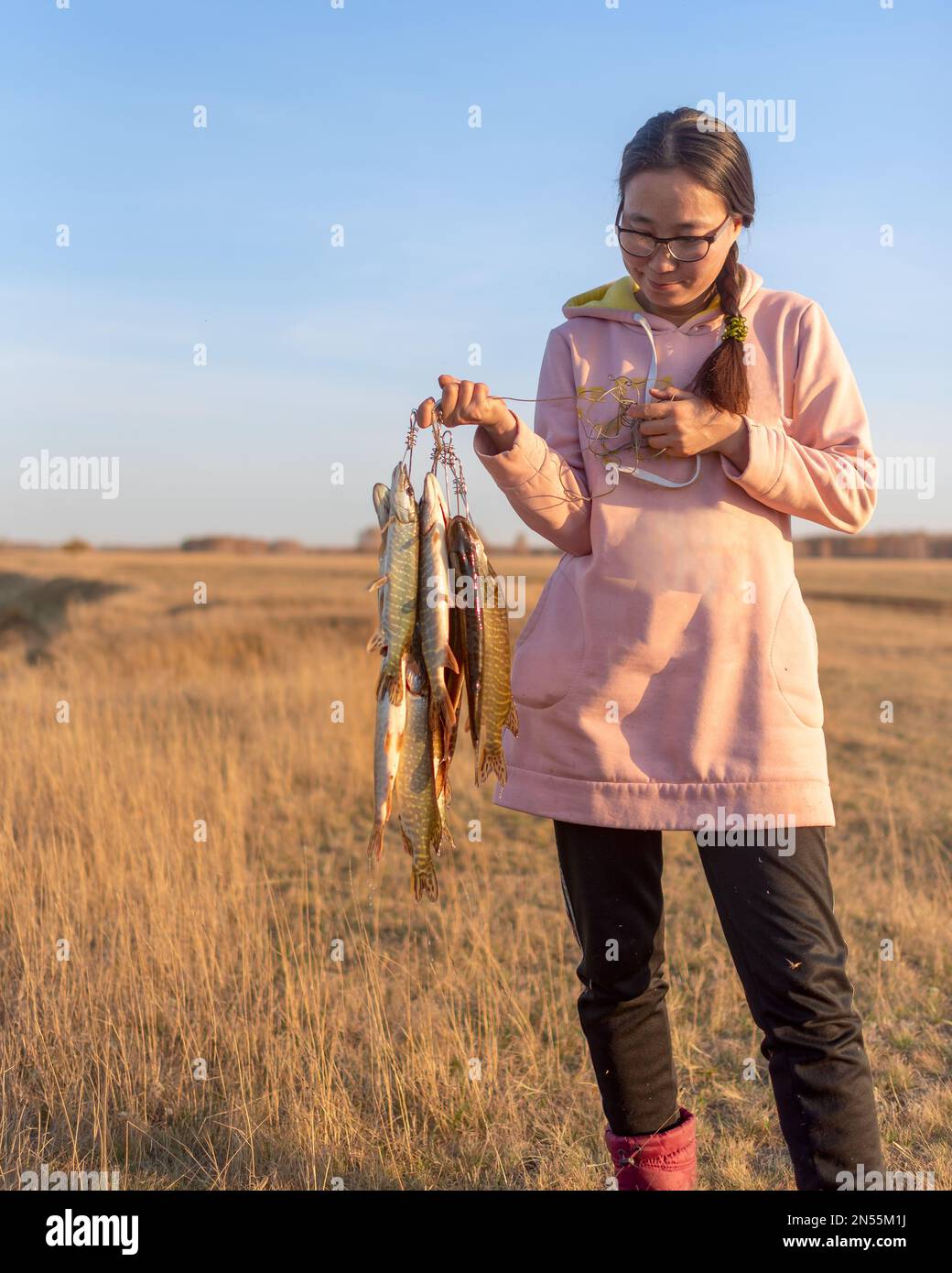 Yakut, junge, fröhliche Anglerin, hält in seinen Händen eine Menge Fische gefangen Hechte hängen auf Fish Stringer auf dem Feld. Stockfoto