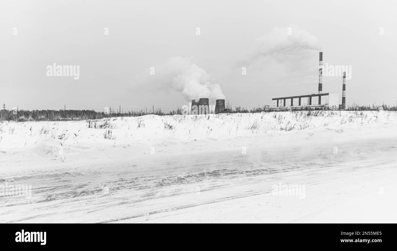 Die Straße mit Schneeverwehungen verläuft vor dem Hintergrund großer Heizkessel eines leistungsstarken Wärmekraftwerks, die im Winter im Himmel Kohlegauch rauchen Stockfoto