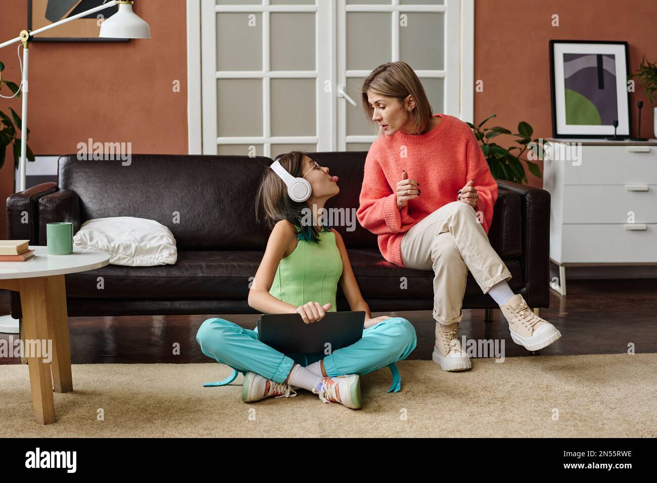 Teenager, die mit dem Laptop auf dem Boden saß und ihre Mutter ärgerte, während sie sich ernsthaft mit ihr im Zimmer zu Hause unterhielt Stockfoto