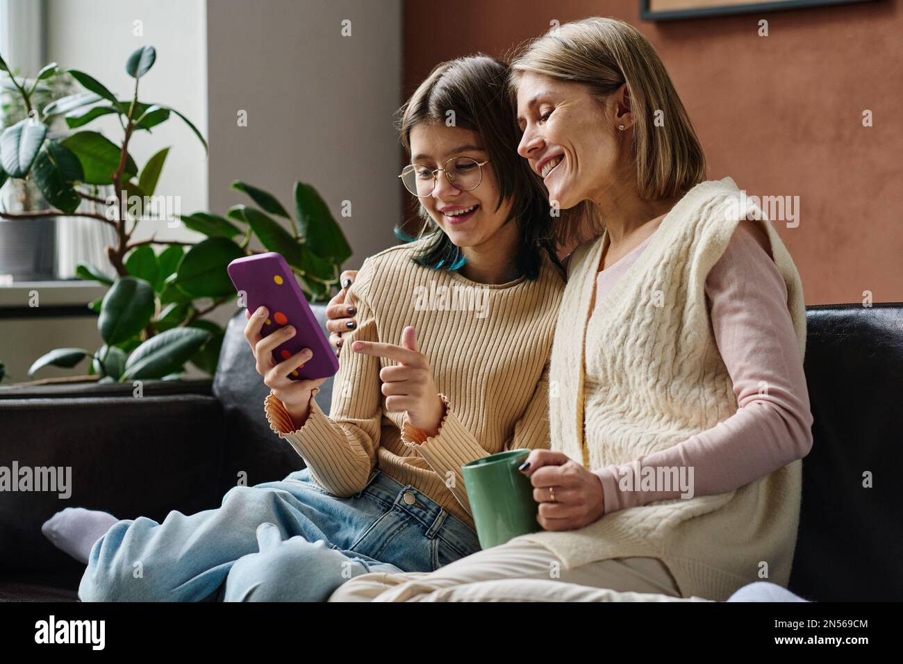 Teenager-Mädchen, die zusammen mit ihrer mutter Fotos auf dem Smartphone anschauen, sie sitzen auf dem Sofa, reden und lächeln Stockfoto